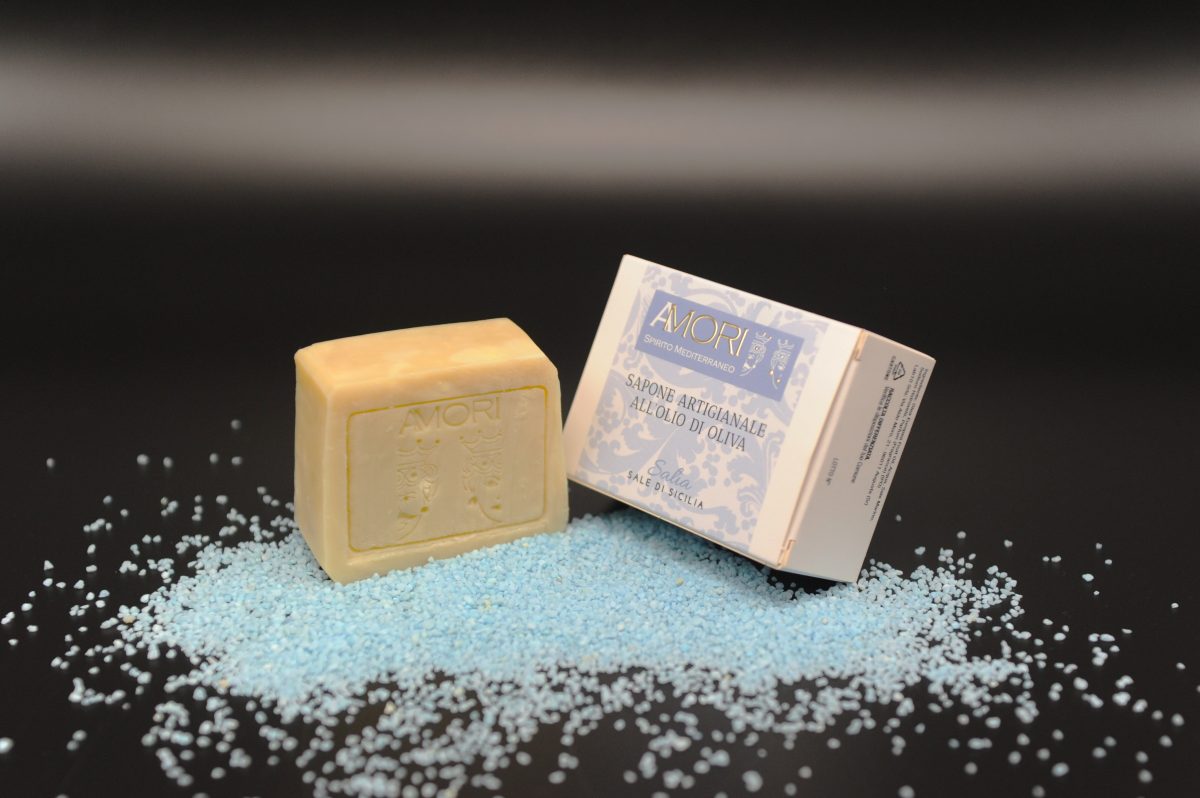 foto sapone al sale marino SALI'A Sapone artigianale all'olio di oliva al profumo di sale marino di Sicilia. Il nostro sapone è fatto secondo un’antica ricetta siciliana del 1800 tramandata di generazione in generazione. Produciamo seguendo il metodo a caldo, per conservare inalterate le proprietà del nostro puro olio di oliva. L’Olivo è il primo albero citato nella storia ed è uno dei più diffusi ed amati in Sicilia. Il suo olio è ricco di acido oleico (circa il 62%), con grandi proprietà nutrienti ed emollienti per la pelle. Il sale marino richiama la purezza del mare, regalando freschezza e benessere. Un gesto semplice, che rispetta la natura e si prende cura di te.